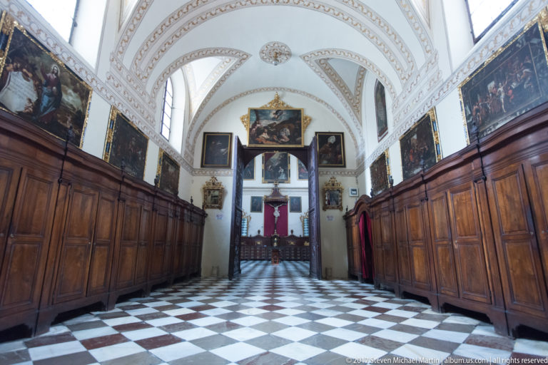 Long wood-paneled cathedral room with religious paintings, ribbed white vault, checkerboard floor, and crucifix at the far end.