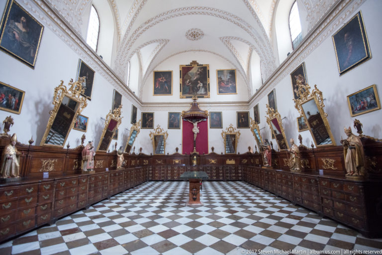 Long cathedral sacristy with wooden drawers, religious paintings and statues, checkerboard floor, and pale vaulted ceiling.