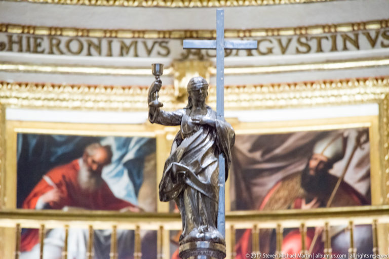 Silver-toned saint figure beside a tall cross, with large religious paintings and gilded moldings behind in Granada Cathedral.