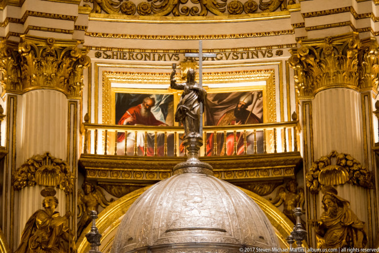 Large domed silver-toned liturgical structure topped by a figure and cross beneath paintings in Granada Cathedral.