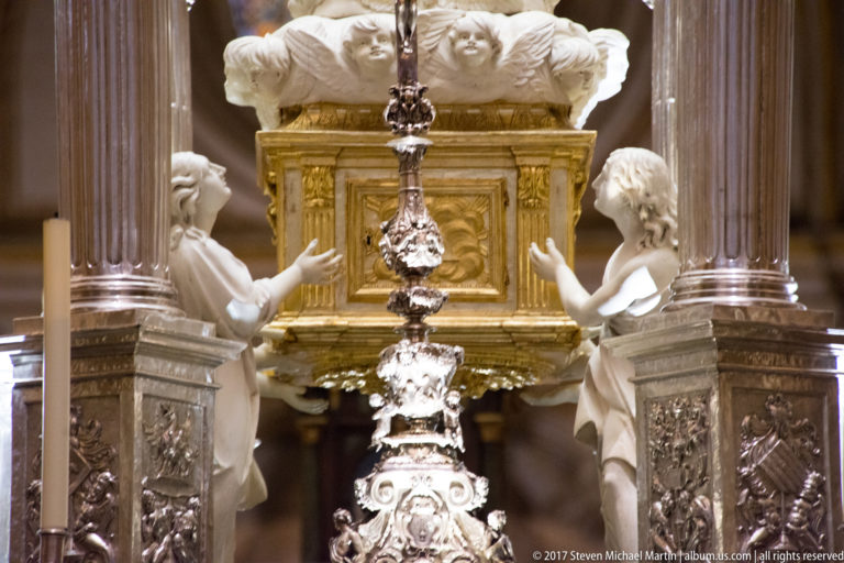 Ornate silver-toned metalwork between dark columns, with two white angel figures and a gilded panel behind.