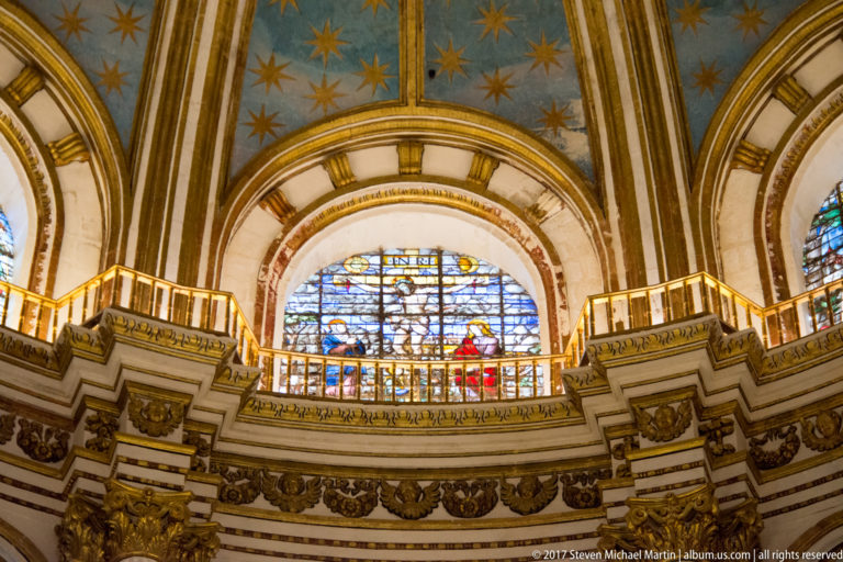 Large stained-glass window above a curved gallery beneath a blue, gold-starred vault in Granada Cathedral.