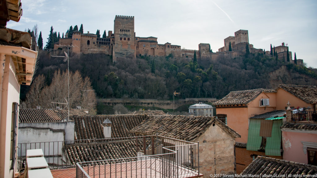 Alhambra walls and towers stretching across a wooded hill above tiled rooftops in Granada.