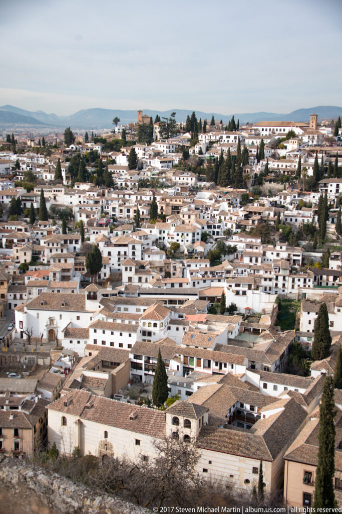 Wide view of low-rise white neighborhoods and tiled roofs spreading across the plain beyond the Alhambra.