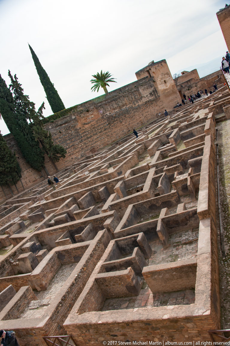Steep view across a dense grid of excavated masonry foundations inside the Alcazaba, with fortress walls and trees above.