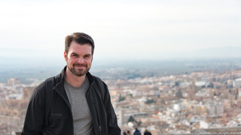 Bearded man in a black jacket posed at an elevated viewpoint with the city of Granada softly blurred behind him.