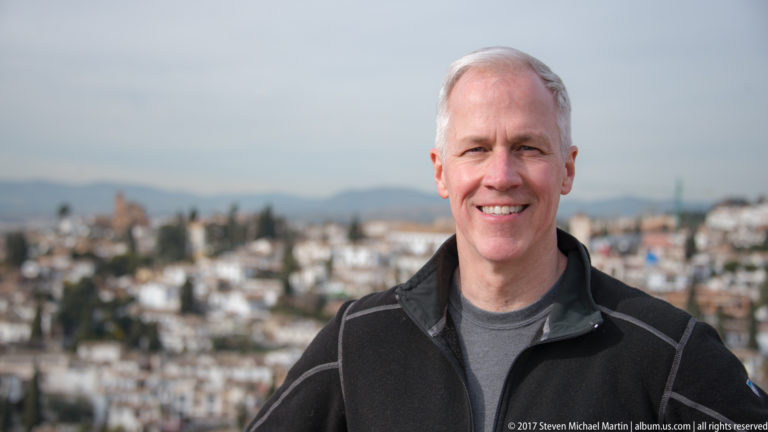 Smiling gray-haired man in a black jacket posed at an elevated viewpoint with Granada blurred in the background.