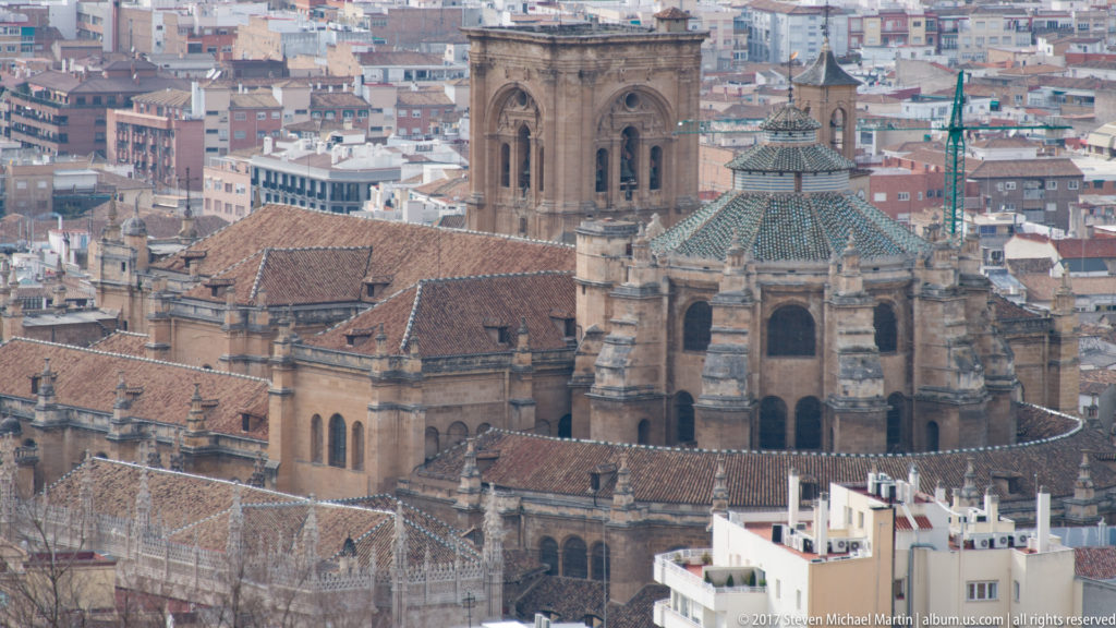 Telephoto view of Granada Cathedral rising above dense city rooftops, with large stone walls, tiled roofs, and projecting chapels.