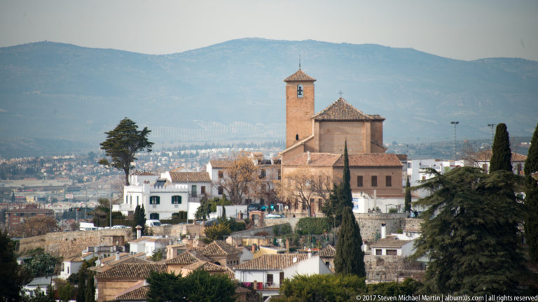 Brick church with square bell tower among white houses and cypress trees on the Albaicín hillside, mountains behind.