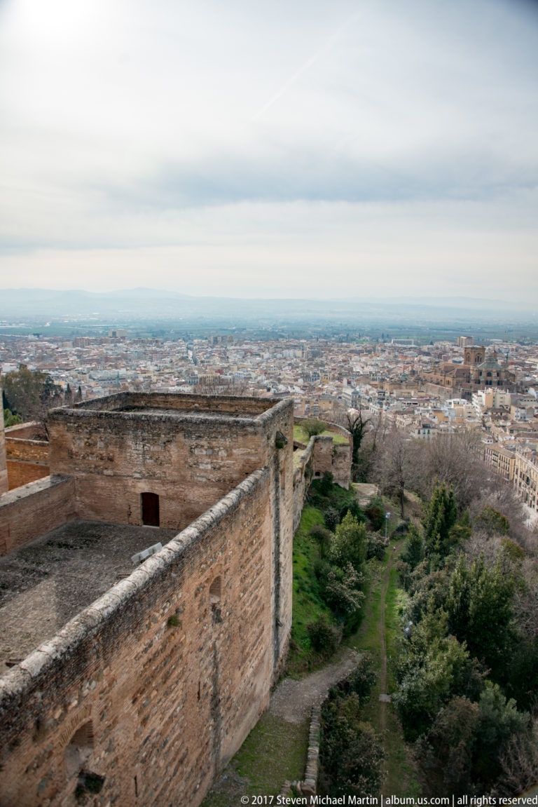 Massive Alhambra wall and flat terrace above green slopes, with Granada extending across the plain in the distance.