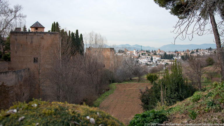 Reddish Alhambra wall beside cultivated terraces and winter trees, with white hillside houses of Granada beyond.