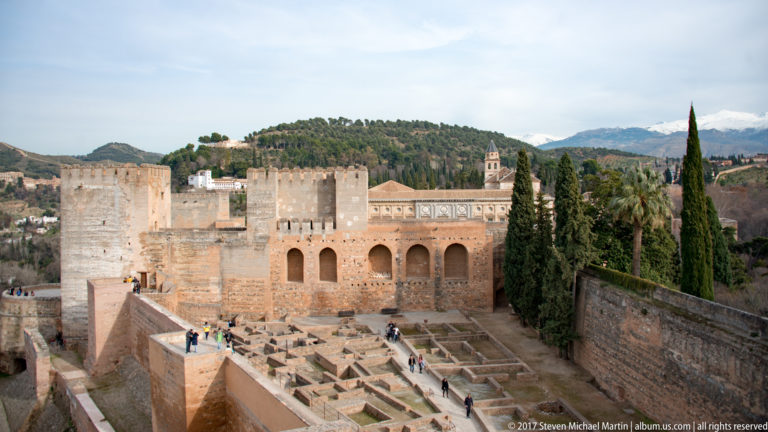 Alcazaba towers above excavated wall foundations and paths, with visitors and cypress trees inside the fortified enclosure.