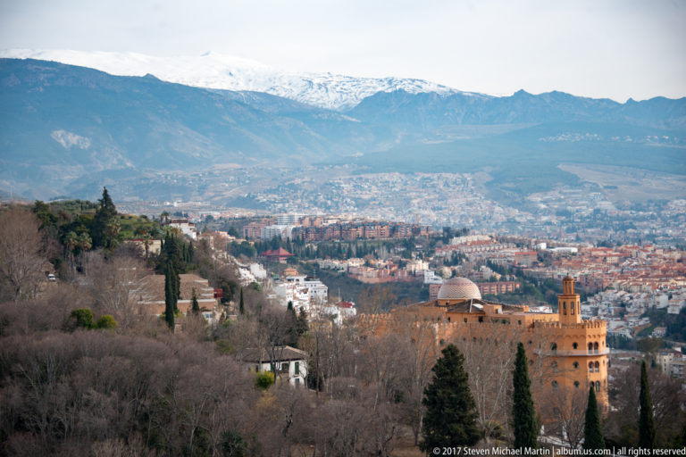 Granada spread below the Alhambra with wooded foreground slopes and snow-covered Sierra Nevada mountains on the horizon.