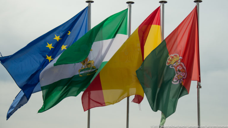 Four flags on tall poles: European Union, Andalusian, Spanish, and a red-and-green local flag with a coat of arms.