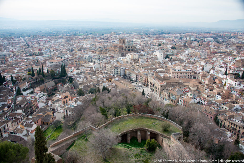 Wide elevated view over Granada with dense rooftops, church towers, foreground walls and terraces, and distant plains.