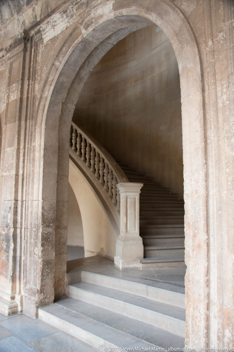 Curving stone staircase with balustrade rising beneath a broad masonry arch inside the Alhambra complex.