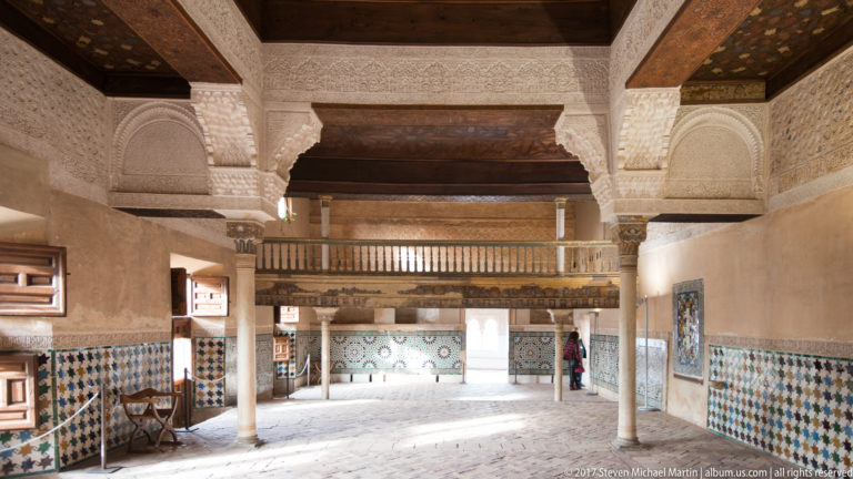 Large palace interior with slender columns, carved arches, patterned tile dados, wooden ceiling beams, and an upper timber gallery.