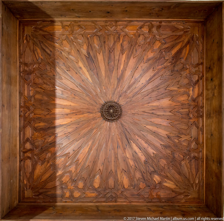 Square wooden ceiling with a small central rosette and radiating geometric starburst pattern in varied tones of timber.