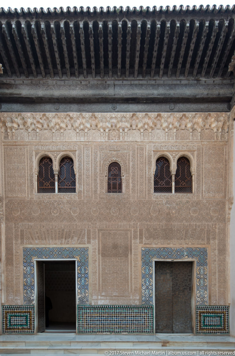 Nasrid façade with paired arched windows, two doorways bordered by tilework, dense carved stucco, and a projecting wooden eave.