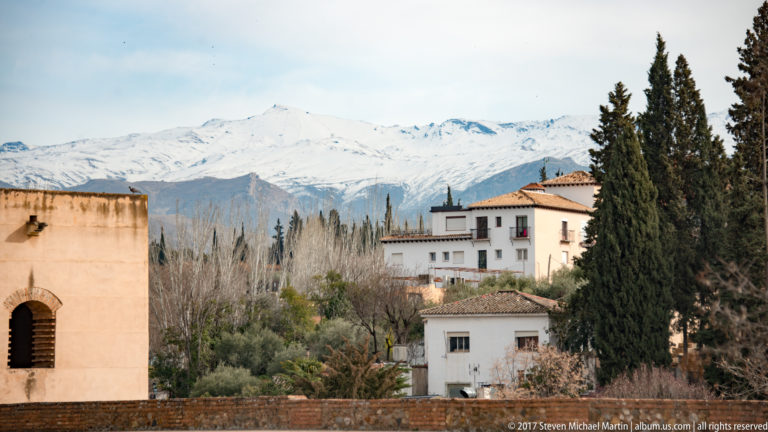 Snow-covered Sierra Nevada mountains behind pale Alhambra buildings, cypress trees, and winter garden vegetation.