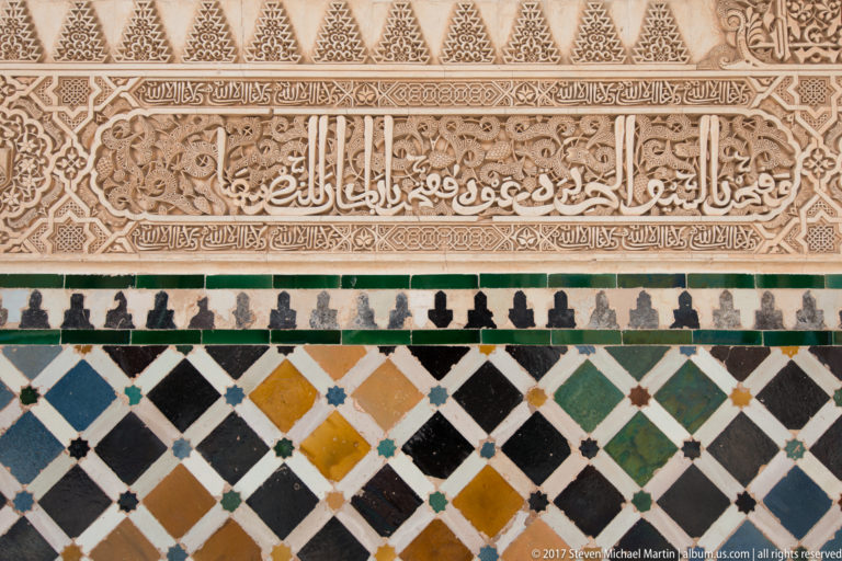 Arabic calligraphy carved in pale stucco above a multicolored geometric tile dado of diamonds and small star motifs.