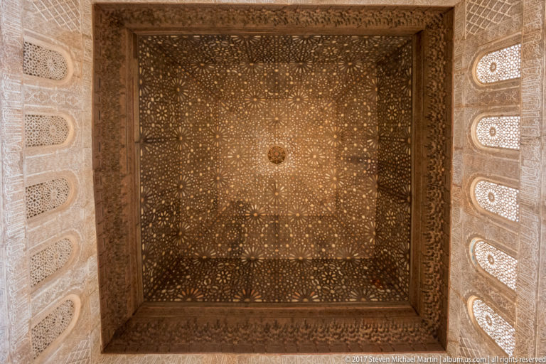 Square dark wooden ceiling with interlocking star patterns and central boss, bordered by carved stucco and small arched windows.