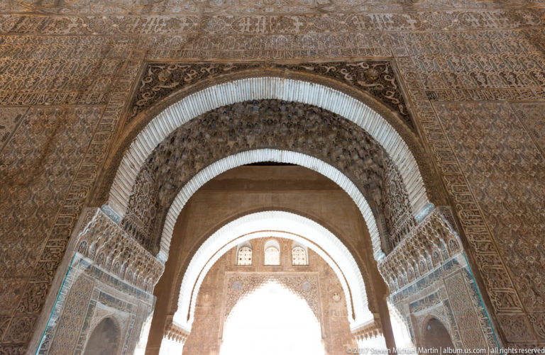 Successive horseshoe arches receding through a richly carved Nasrid interior toward bright daylight.