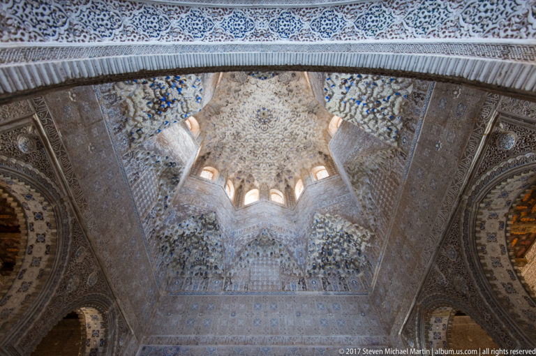 Star-shaped muqarnas dome with small windows viewed past a dark carved stucco arch in the foreground.