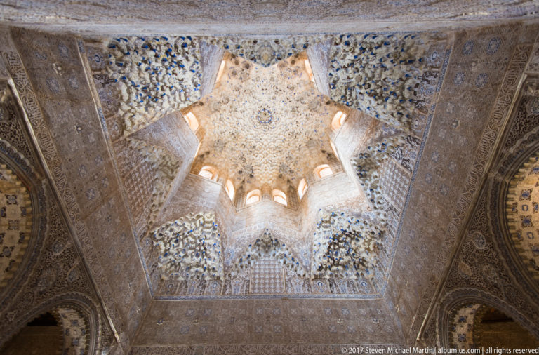 Centered upward view of a star-shaped muqarnas dome with radiating plaster cells and small windows around its base.