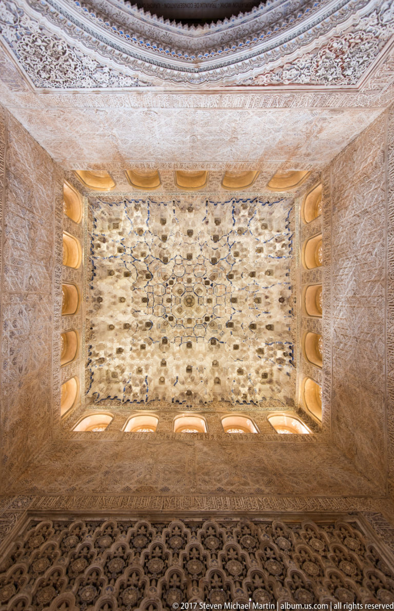 Upward view of an intricate pale muqarnas vault surrounded by carved stucco and small glowing windows.