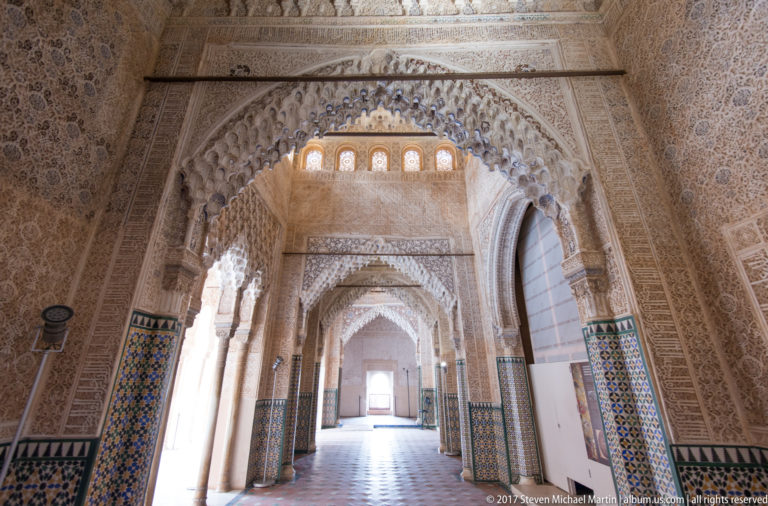 Long Nasrid corridor seen through a muqarnas-edged arch, with tiled lower walls and successive decorated doorways.