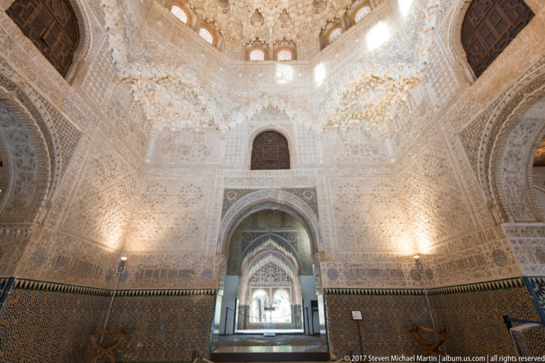 Nasrid hall with carved stucco walls, dark patterned tile dado, and aligned arched doorways beneath a muqarnas vault.