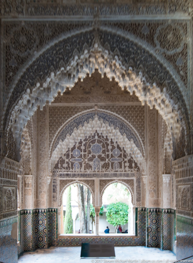 Layered carved Nasrid arches with patterned tile dados opening toward a bright garden and trees beyond.