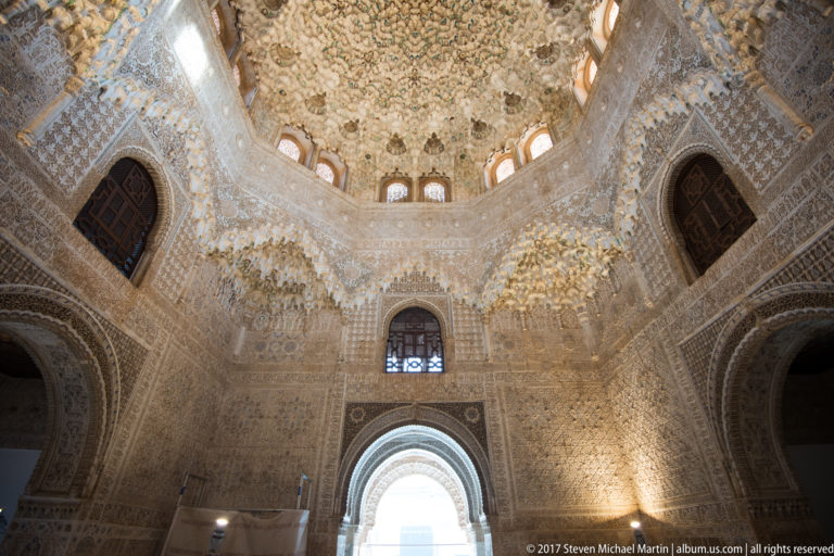High Nasrid chamber with a luminous muqarnas dome, carved stucco walls, upper windows, and a bright arched doorway.