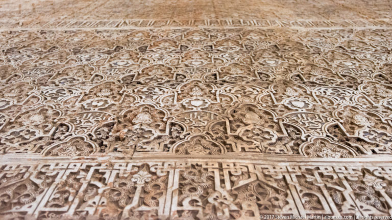 Close view of pale carved Nasrid stucco with dense arabesques, geometric motifs, and a horizontal Arabic inscription band.