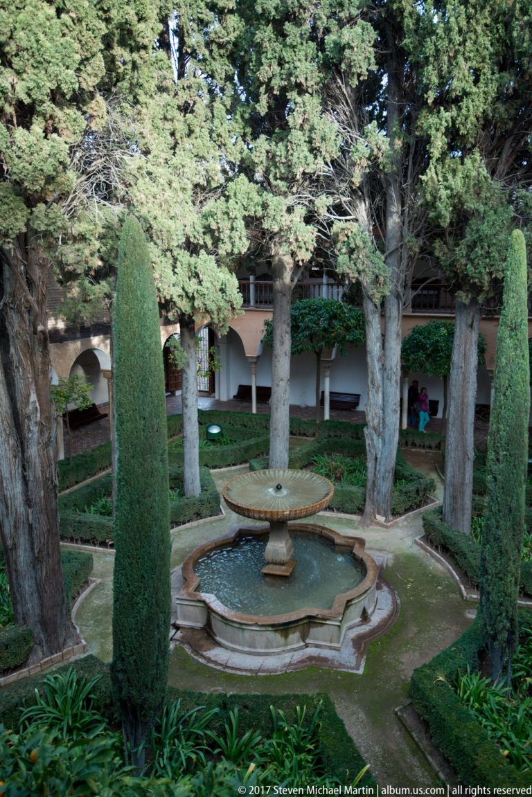 Tiered stone fountain centered among cypress trees, mature plantings, geometric beds, and arcaded walls at the Generalife.