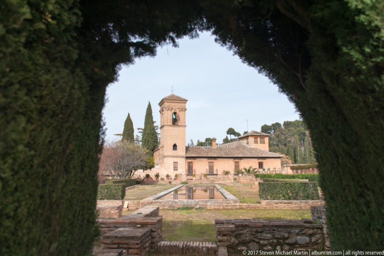Historic pavilion and terraced garden seen through a dark opening cut into tall cypress hedges.