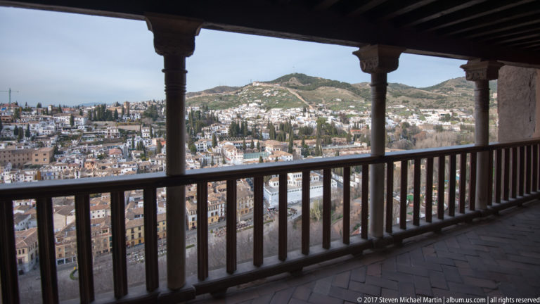 Granada and surrounding hills viewed through a dark wooden gallery with posts, beams, and a balustrade.