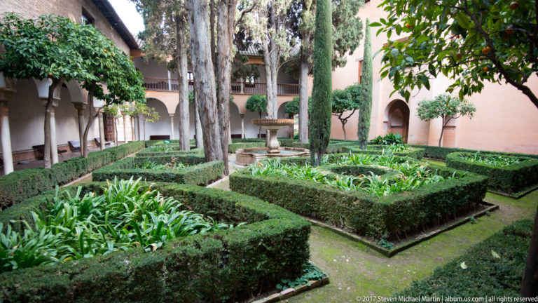 Enclosed Generalife garden with geometric hedges, tall cypress trees, a stone fountain, and arcaded walls.