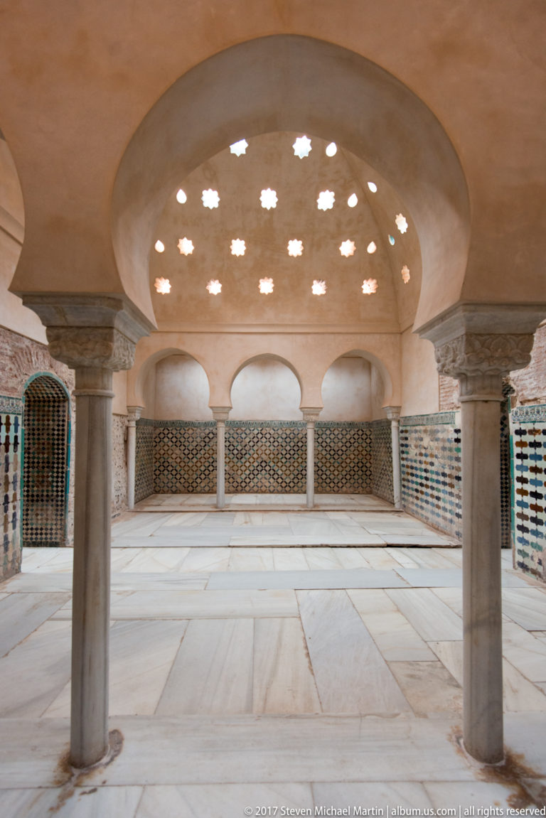 Low chamber with stone columns, patterned tilework, grilles, and a shallow ceiling pierced by many star-shaped openings.