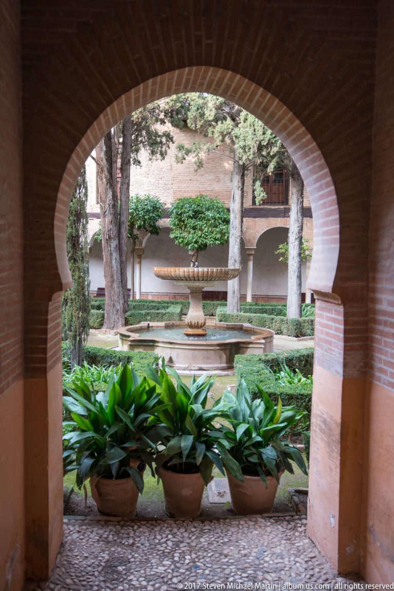 Brick arch framing a Generalife garden with a tiered stone fountain, potted plants, mature trees, and arcaded walls.