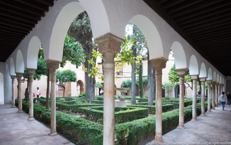 Whitewashed Generalife arcade with slender stone columns enclosing geometric hedges and mature garden trees.