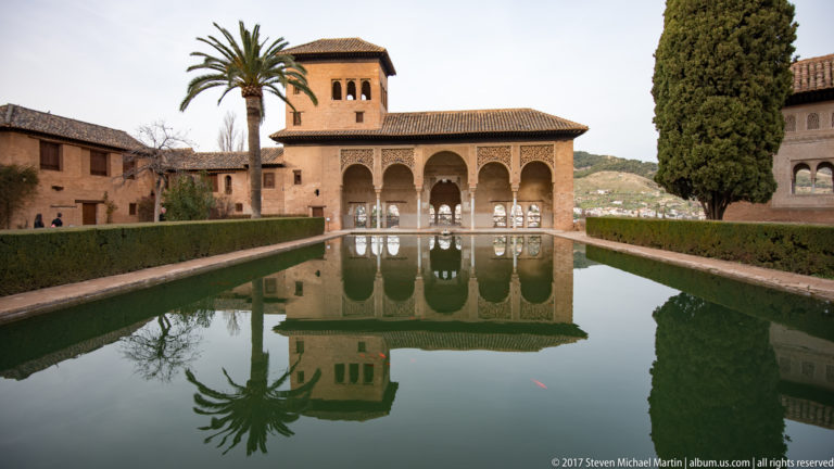 Arcaded Generalife pavilion and tiled roof reflected in a long rectangular pool bordered by clipped hedges and trees.