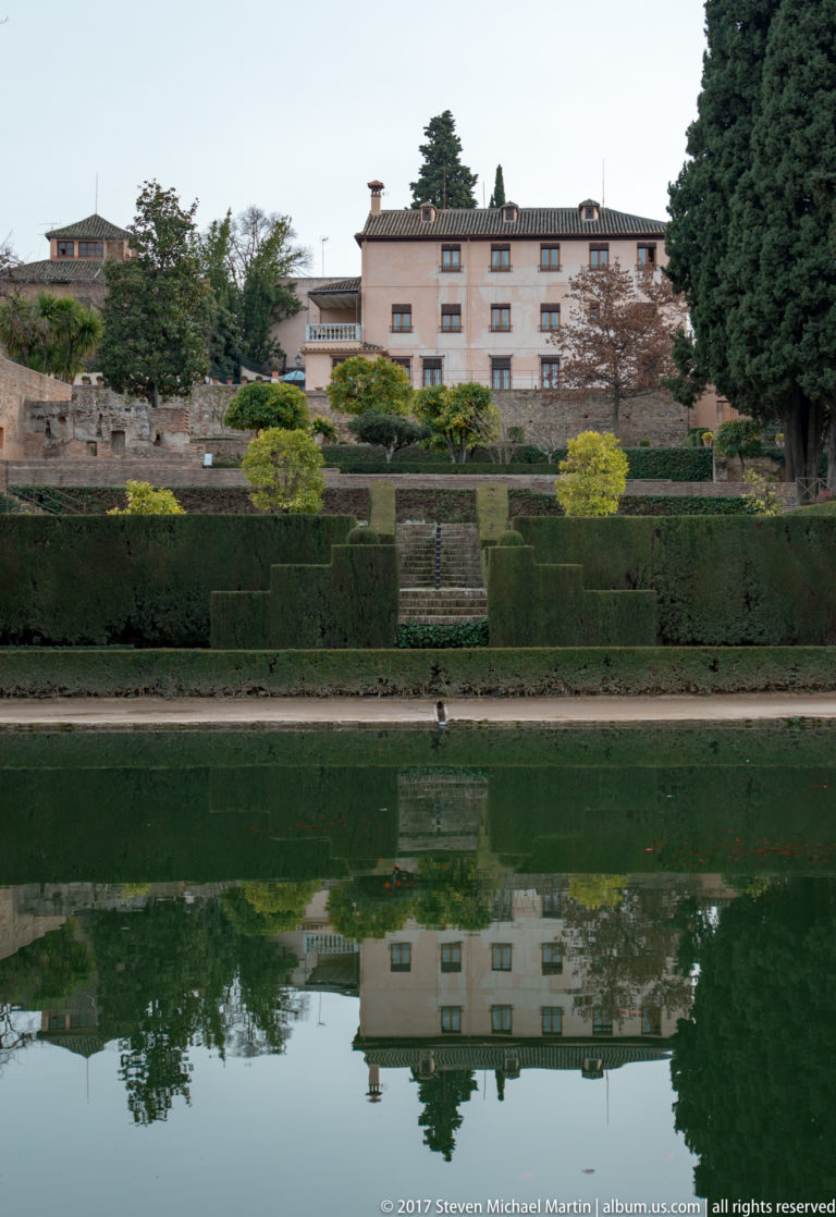 Long reflecting pool mirroring clipped hedges, terraced gardens, trees, and a pale building at the Generalife.