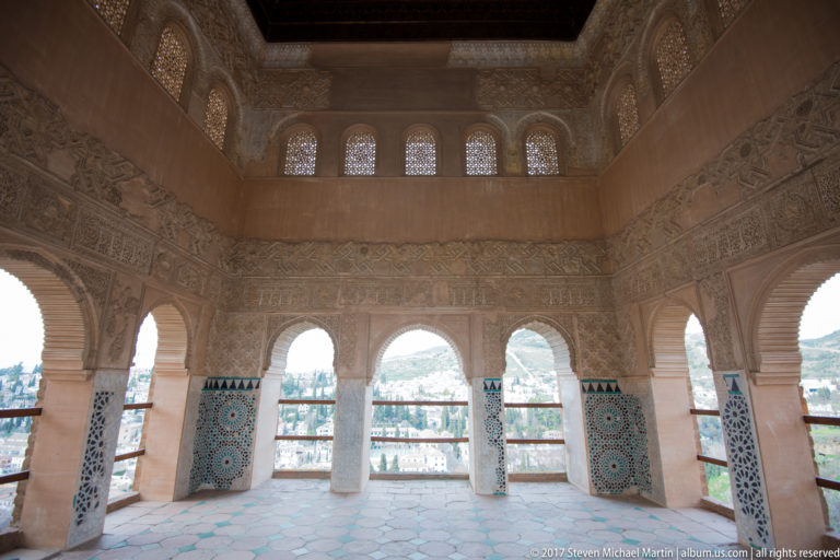 Open Generalife arcade with carved plaster, patterned tile dados, lattice windows, and views toward Granada.
