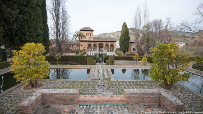 Formal Generalife garden with rectangular reflecting pools, clipped hedges, cypress trees, and an arcaded pavilion beyond.