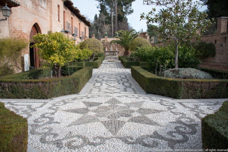Black-and-white pebble-mosaic garden path bordered by clipped hedges, citrus trees, palms, and brick garden walls.