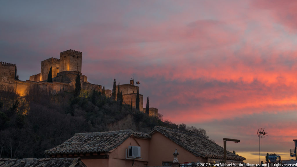 Illuminated Alhambra towers and walls on a wooded ridge beneath a vivid red and violet sunset sky.