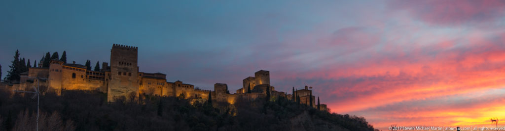 Wide sunset view of the illuminated Alhambra on its ridge, with towers and crenellated walls beneath pink and orange clouds.