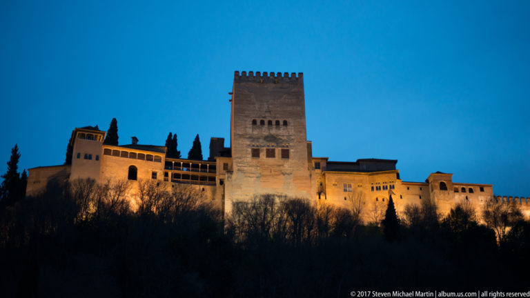 Illuminated square Comares Tower and adjoining Alhambra buildings above a dark wooded slope under a deep blue evening sky.